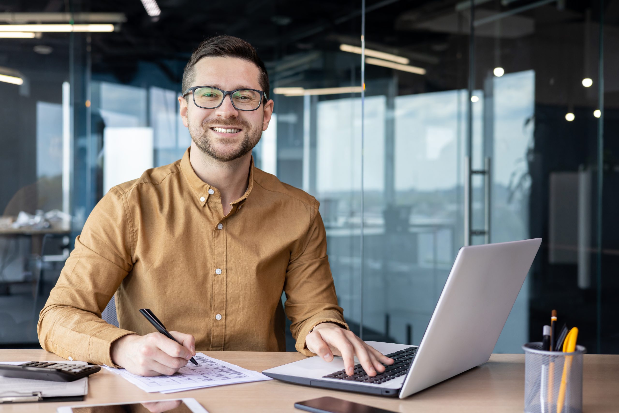 Portrait of young businessman in shirt, man smiling and looking at camera at workplace inside office, accountant with calculator behind paper work signing contracts and financial reports